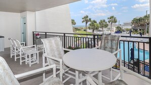 Un balcon avec des chaises blanches et une table, donnant sur une piscine et des palmiers.