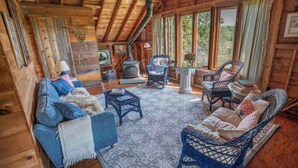A cozy log cabin interior with a sofa, wicker chairs, a fireplace, and large windows. - Barn at Blue Hill Peninsula Historic farm w/Organic Meadows. Pet friendly (Brooksville)