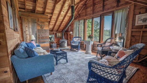 A cozy cabin interior with a blue sofa, wicker chairs, a fireplace, and large windows offering a view of the outdoors. - Barn at Blue Hill Peninsula Historic farm w/Organic Meadows. Pet friendly (Brooksville)