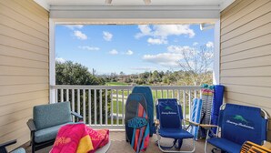 A balcony with a view of a golf course, a surfboard, and several beach chairs. - Luxury Golf-View Condo • Barefoot Resort • Resort Pools • Premium Beach Gear (North Myrtle Beach)