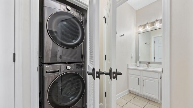 A laundry room with two stacked front-loading washing machines, a white door, and a bathroom with a sink and mirror. - Luxury Golf-View Condo • Barefoot Resort • Resort Pools • Premium Beach Gear (North Myrtle Beach)