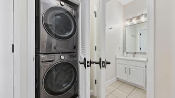 Une buanderie équipée de deux machines à laver à chargement frontal superposées, d’une porte blanche et d’une salle de bain avec un lavabo et un miroir.