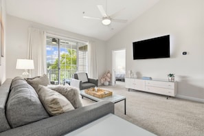 A modern living room with a grey sofa, a coffee table, a TV mounted on the wall, and a sliding glass door leading to a balcony.