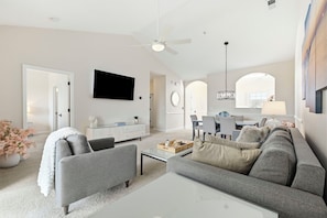 A modern living room with a flat-screen TV, a ceiling fan, and a dining area with a chandelier.