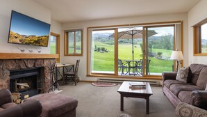 A living room with a fireplace, large windows, a television, and a view of a green landscape. - Villas at Snowmass Club - 1516 ~ Snowmass Club Access ~ (Snowmass Village)