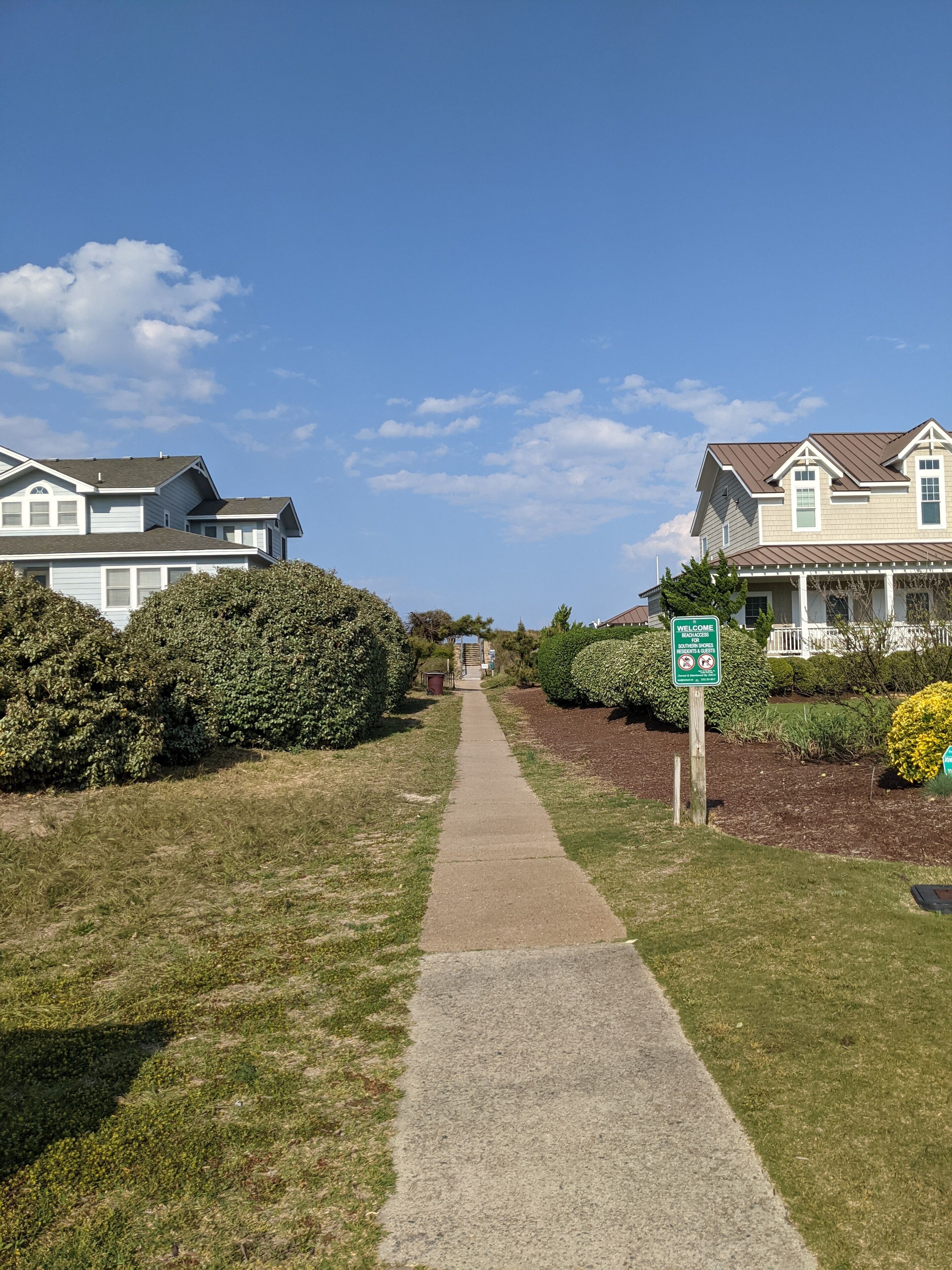 Un chemin mène à une rangée de maisons, agrémentée d’une pancarte « Bienvenue », et bordé de pelouses et de haies bien entretenues.