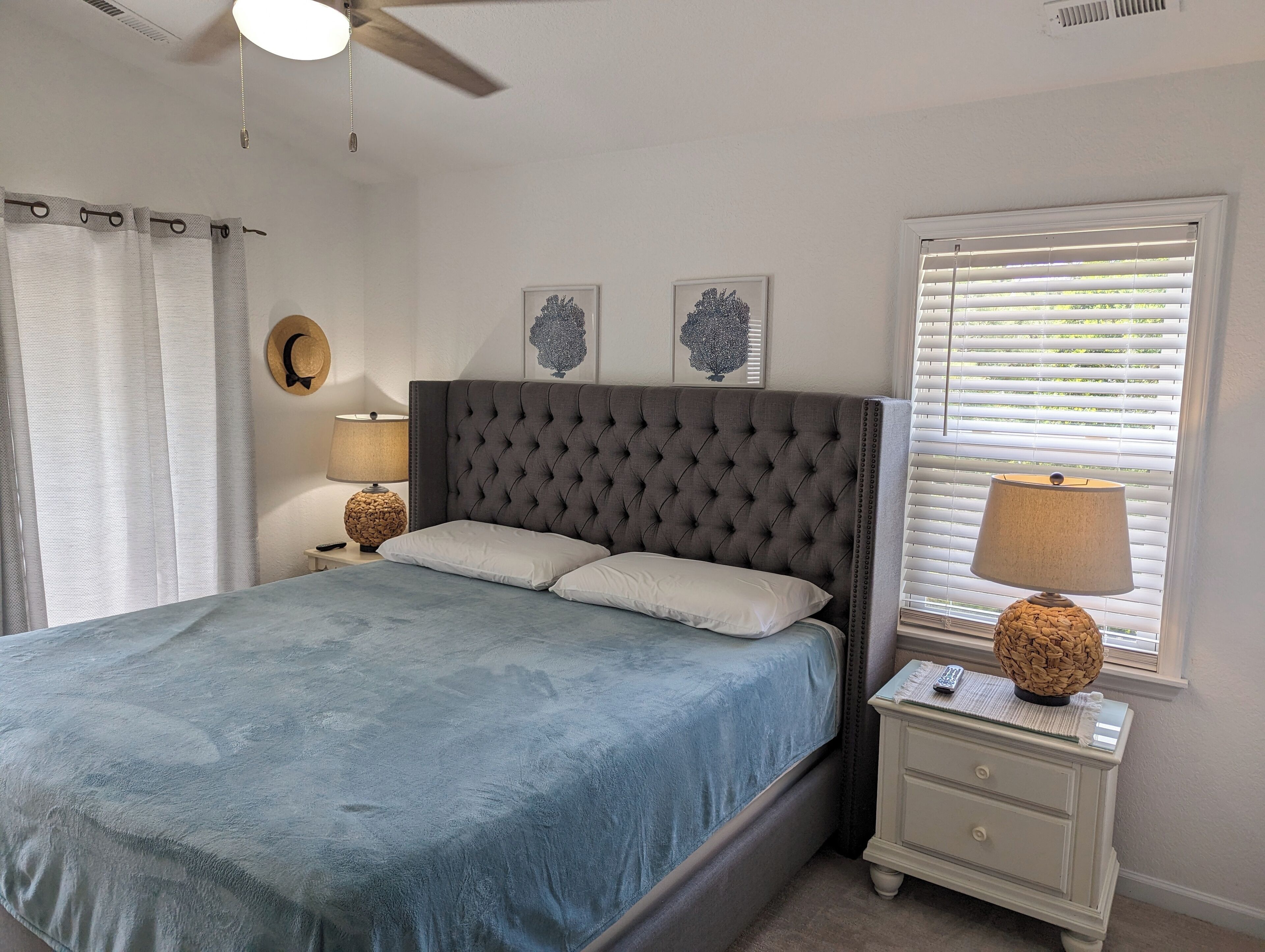 A bedroom with a tufted headboard, a bed with a blue bedspread, a nightstand with a lamp, and a window with blinds.