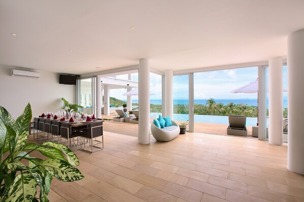 A spacious living area with a dining table set for a meal, a comfortable seating area with a view of the ocean, and large windows allowing natural light.