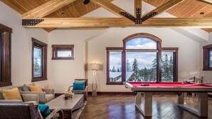 Ein großzügiges Wohnzimmer mit Billardtisch, bequemen Sitzgelegenheiten, großen Fenstern mit Blick auf Bäume und Holzböden.