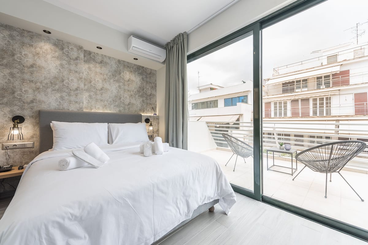 A modern hotel room with a large bed, a view of the city through floor-to-ceiling windows, and a balcony with a chair.