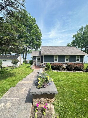 A two-story house with a stone pathway leading to the front door, surrounded by a well-maintained lawn and flowering plants. - WOLCOTT NY  Waterfront cottage rental on Port Bay seasonal available 2026 (Port Bay - Wolcott)