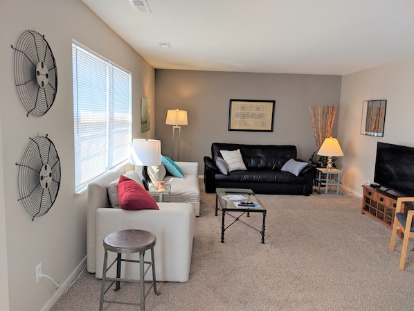 A living room with a black leather sofa, a white armchair, a glass coffee table, and a TV stand with a flat-screen TV.