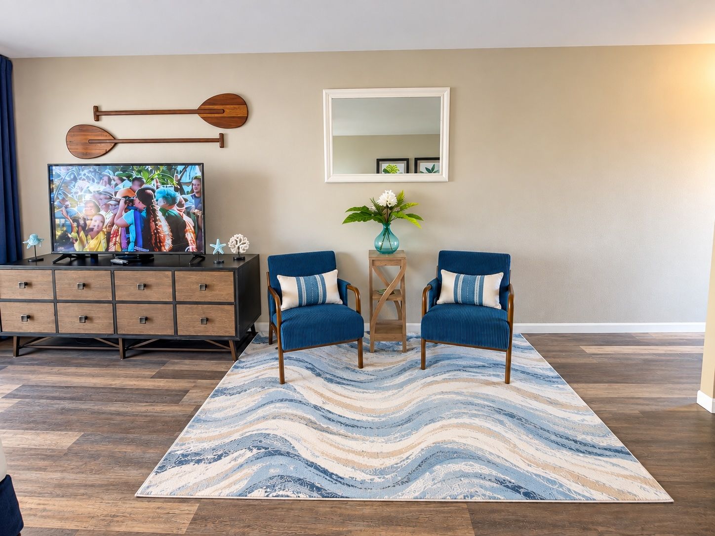Ein Wohnzimmer mit Fernseher, zwei blauen Sesseln, einem Holzschrank, einem Spiegel und einem gemusterten Teppich.