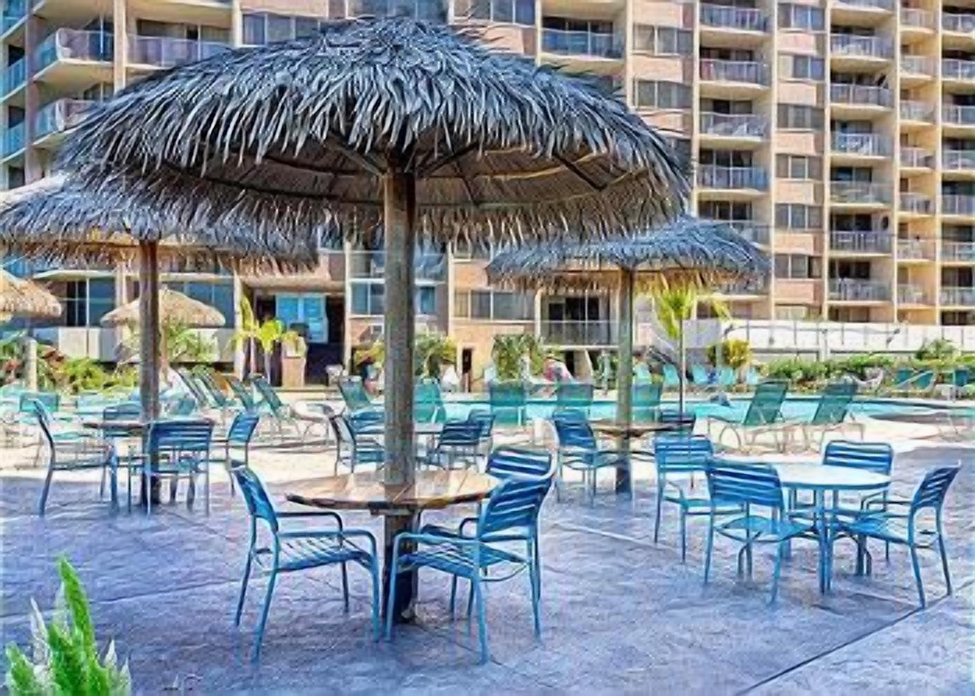 A poolside area with lounge chairs and umbrellas, a building in the background, and a clear sky.