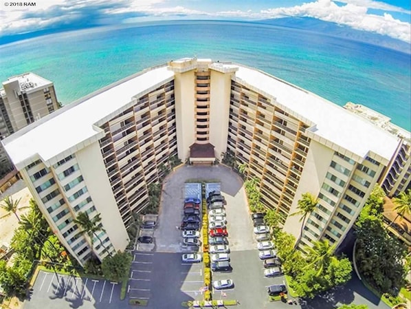 A high-rise hotel with a central courtyard parking area, surrounded by palm trees and ocean views.