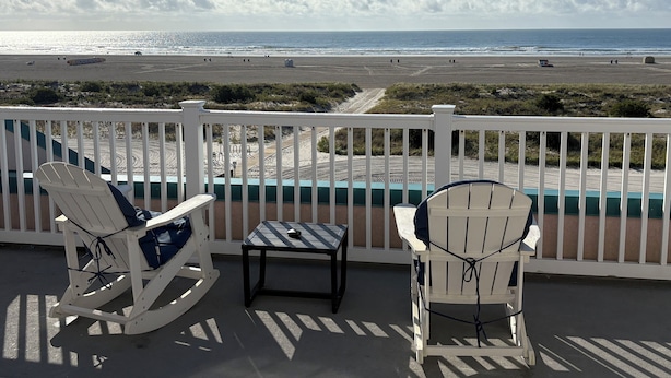Een balkon met twee Adirondack stoelen en een kleine tafel, met uitzicht op een strand en een helder zicht op de oceaan en de verre kustlijn.