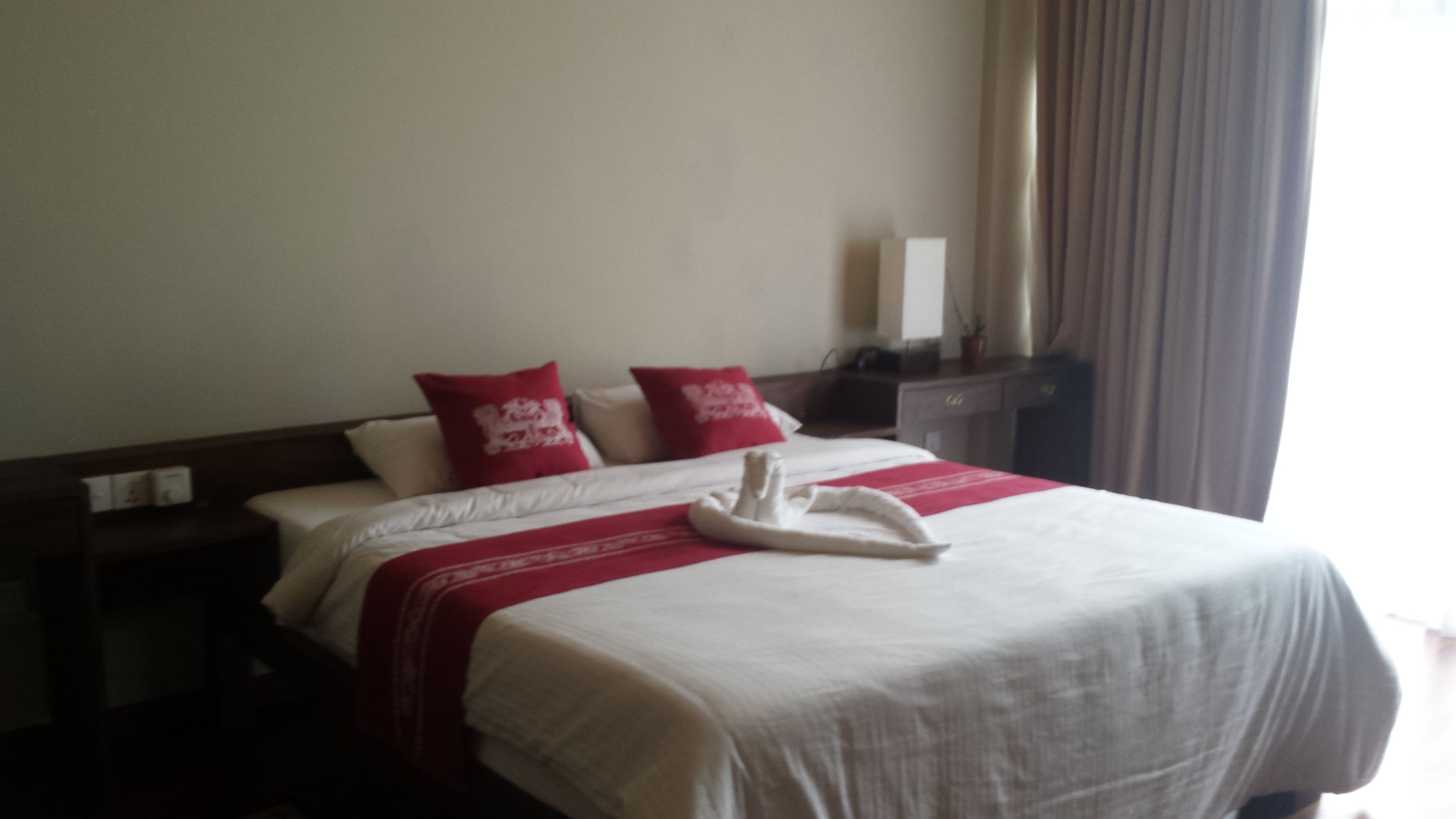 A hotel room with a neatly made bed, two red pillows, a white towel, and a nightstand with a lamp.