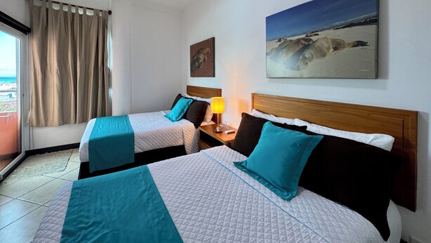 A hotel room with two beds, a window with curtains, a painting on the wall, and a lamp on the nightstand. - The Oceanview two-bedroom with balcony (Puerto Ayora, Santa Cruz Island)