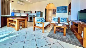 A living room with a wooden coffee table, two sofas with blue cushions, a TV stand, and a kitchen area with a refrigerator and a water dispenser. - The Oceanview two-bedroom with balcony (Puerto Ayora, Santa Cruz Island)