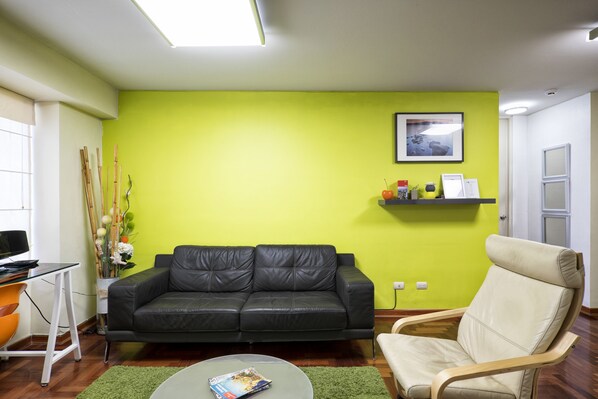 Een woonkamer met een zwarte leren bank, een beige fauteuil, een glazen salontafel en een heldergroene muur.