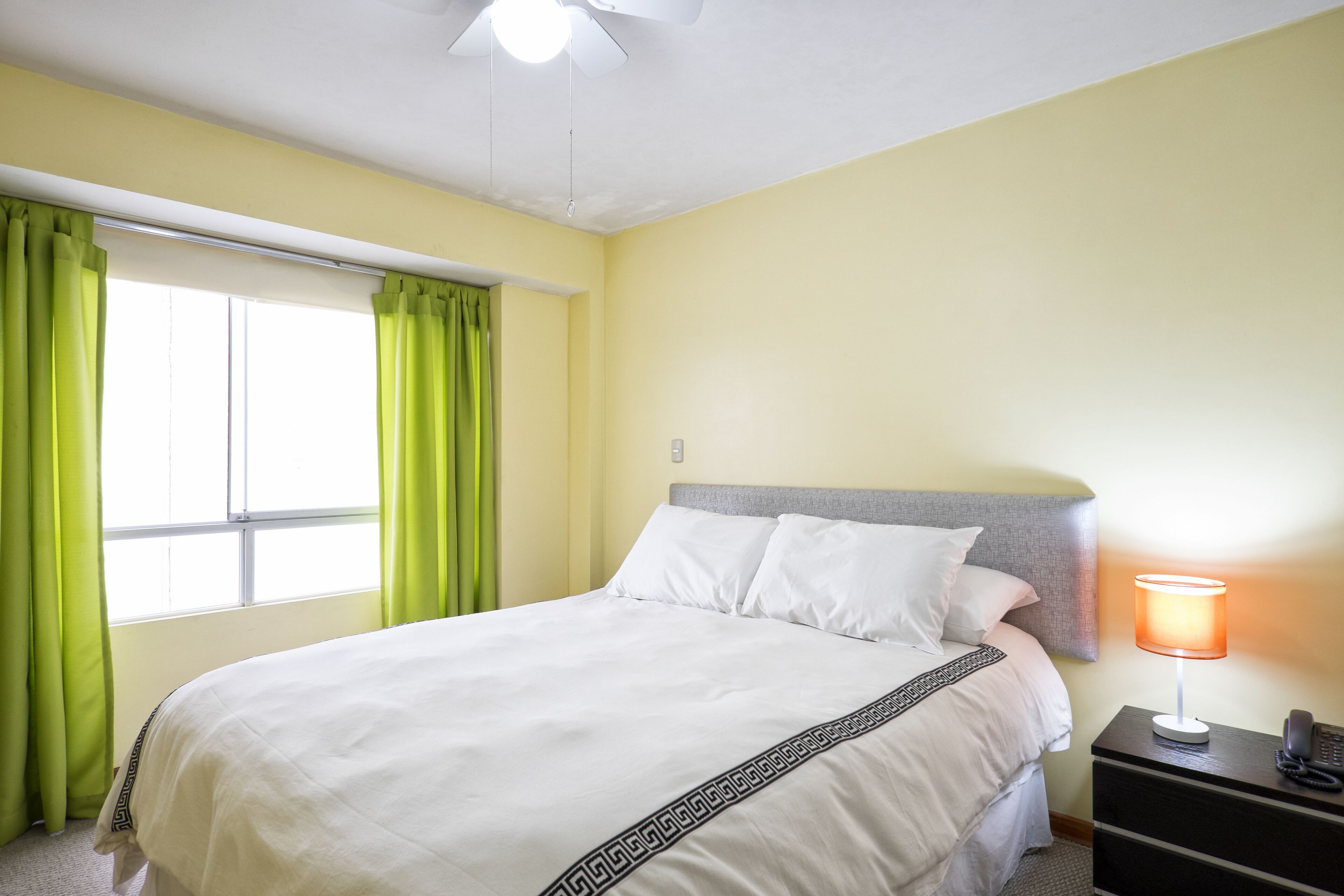A neatly made bed with white linens and a black geometric border, a bedside lamp, and a nightstand with a telephone.