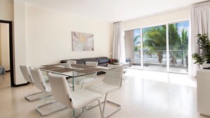 A modern living room with a glass dining table, white chairs, a grey sofa, and a large window with curtains.