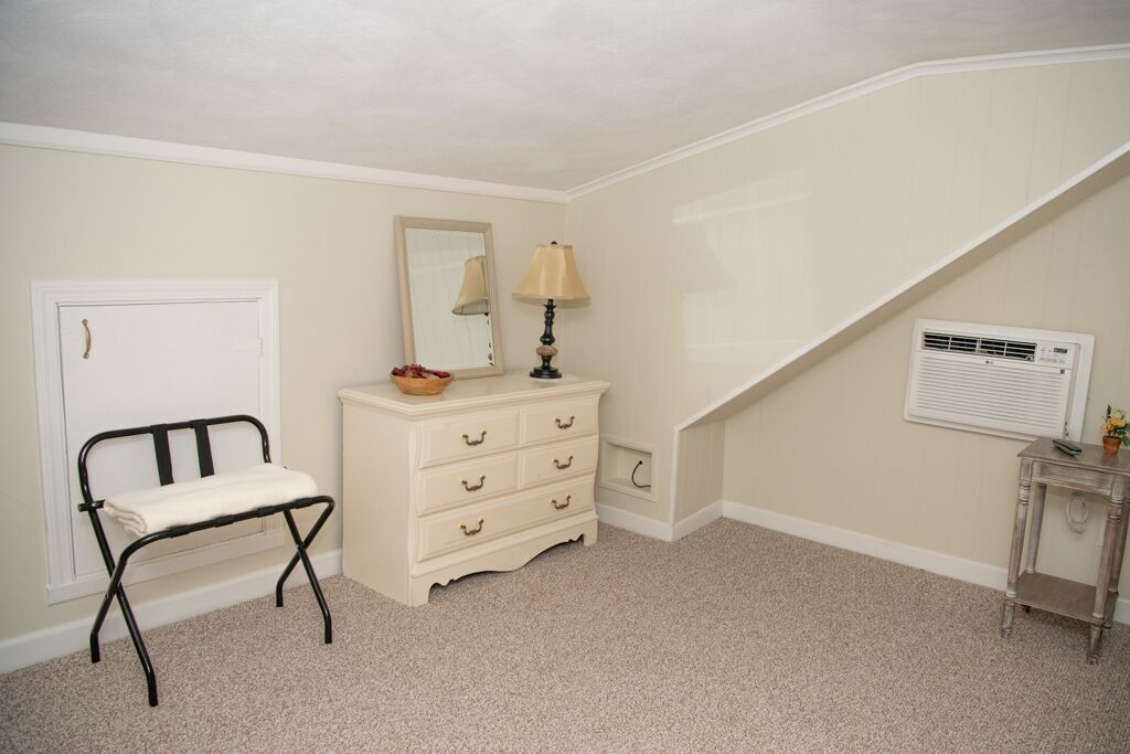 A small, neatly organized room with a white door, a bench, a dresser with drawers, a mirror, a lamp, and an air conditioning unit.