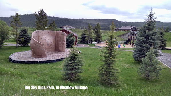 Un parque infantil con estructuras para escalar y un tobogán, rodeado de árboles y casas en Meadow Village.