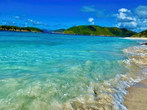 A beach with clear turquoise water, a sandy shore, and green hills in the background.