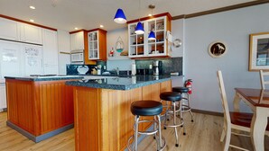 A kitchen with wooden cabinetry, a central island with stools, blue pendant lights, and a fire extinguisher. - OCEAN FRONT: SEASCAPE CONDO:  Seaside Vacation Homes, Inc Managed  (Seaside)