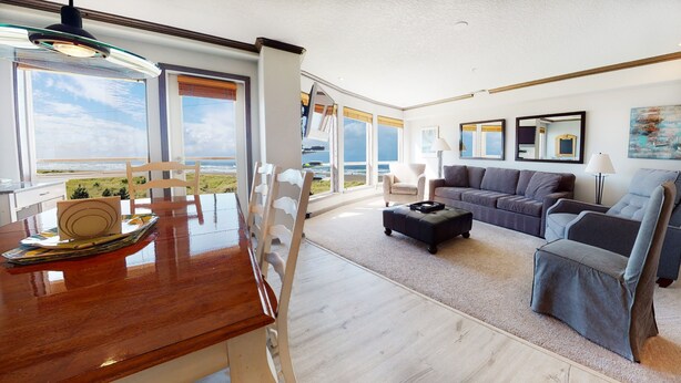 A spacious living room with a wooden dining table, a sofa, and a large window offering a view of the sea. - OCEAN FRONT: SEASCAPE CONDO:  Seaside Vacation Homes, Inc Managed  (Seaside)