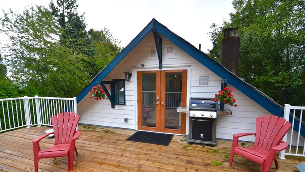 En liten, enplansstuga med ett brant tak och en veranda. På verandan står två Adirondack-stolar, en grill och en dörr med ett glasparti.