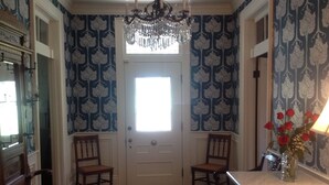 A room with a chandelier, two wooden chairs, a door with a window, and a vase with red roses on a counter.