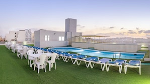 Ein Poolbereich auf dem Dach mit mehreren Liegestühlen und Tischen, einem Schwimmbecken und einer Stadtansicht im Hintergrund.