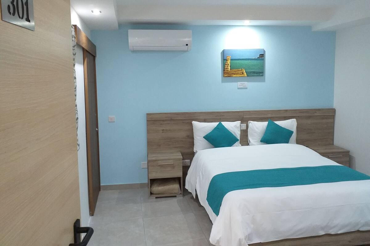 Une chambre à coucher comprenant un lit, des tables de chevet, un climatiseur fixé au mur et un tableau représentant un paysage côtier.
