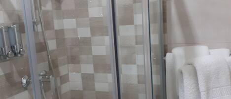 Ducha con cerramiento de vidrio, pared de azulejos a cuadros, cabezal de ducha manual y toallero con toallas blancas.