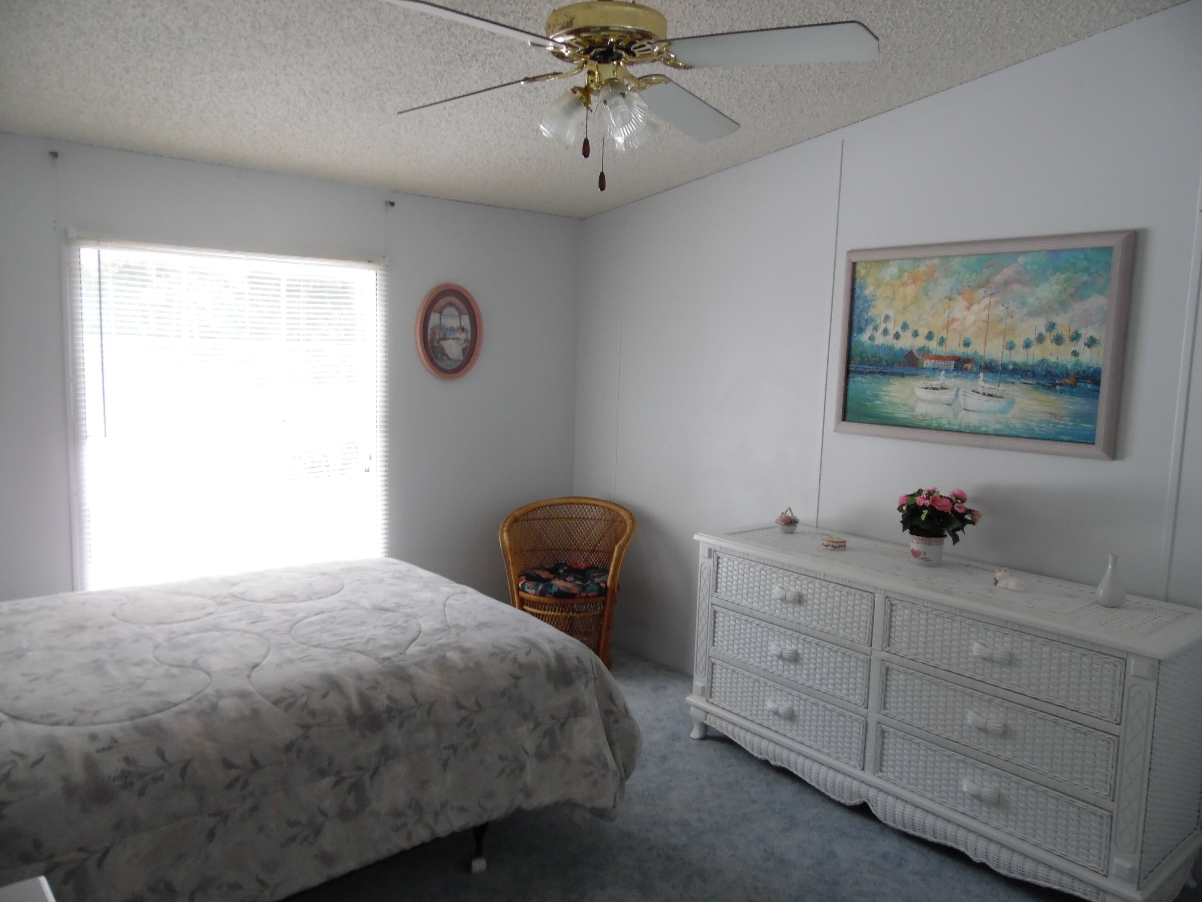 Une chambre à coucher comprenant un lit, une chaise, une commode, un ventilateur de plafond, une fenêtre et un tableau accroché au mur.