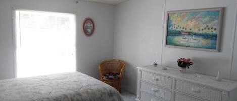Une chambre à coucher comprenant un lit, une chaise, une commode, un ventilateur de plafond, une fenêtre et un tableau accroché au mur.