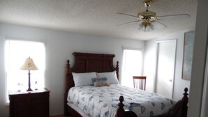 Un dormitorio con una cama de madera, una mesita de noche con lámpara, un ventilador de techo y una ventana con persianas.
