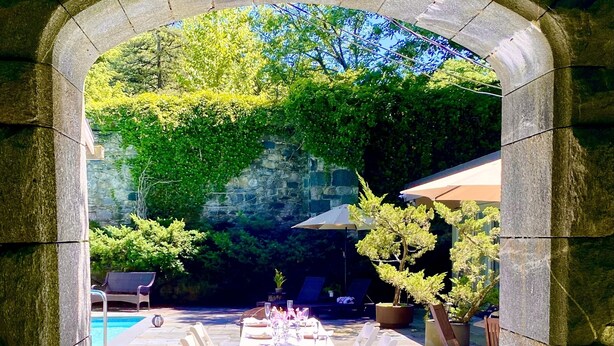 Ein Essbereich im Freien mit einem Steingewölbe, gedeckten Tischen und Blick auf einen Pool und Grünflächen.