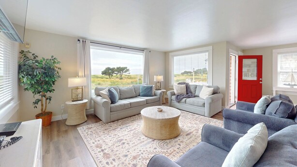 A spacious living room with two sofas, a coffee table, and a large window offering a view of a field and tree. - OCEAN FRONT: ONCE UPON A TIDE:  Seaside Vacation Homes, Inc Managed  (Seaside)
