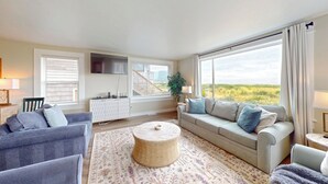 A modern living room with a sofa, armchairs, a round coffee table, and large windows offering a view of the outdoors. - OCEAN FRONT: ONCE UPON A TIDE:  Seaside Vacation Homes, Inc Managed  (Seaside)