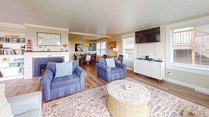 A living room with two sofas, a fireplace, a dining area, and a TV mounted on the wall. - OCEAN FRONT: ONCE UPON A TIDE:  Seaside Vacation Homes, Inc Managed  (Seaside)