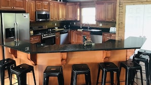 A kitchen with wooden cabinets, a stainless steel refrigerator, a microwave, a stove, and a sink. There are four black bar stools around the island. - Pirates Cove 9 Bed/9 Bath,  large hot tub,  private fire pit, double dock  (La Follette)