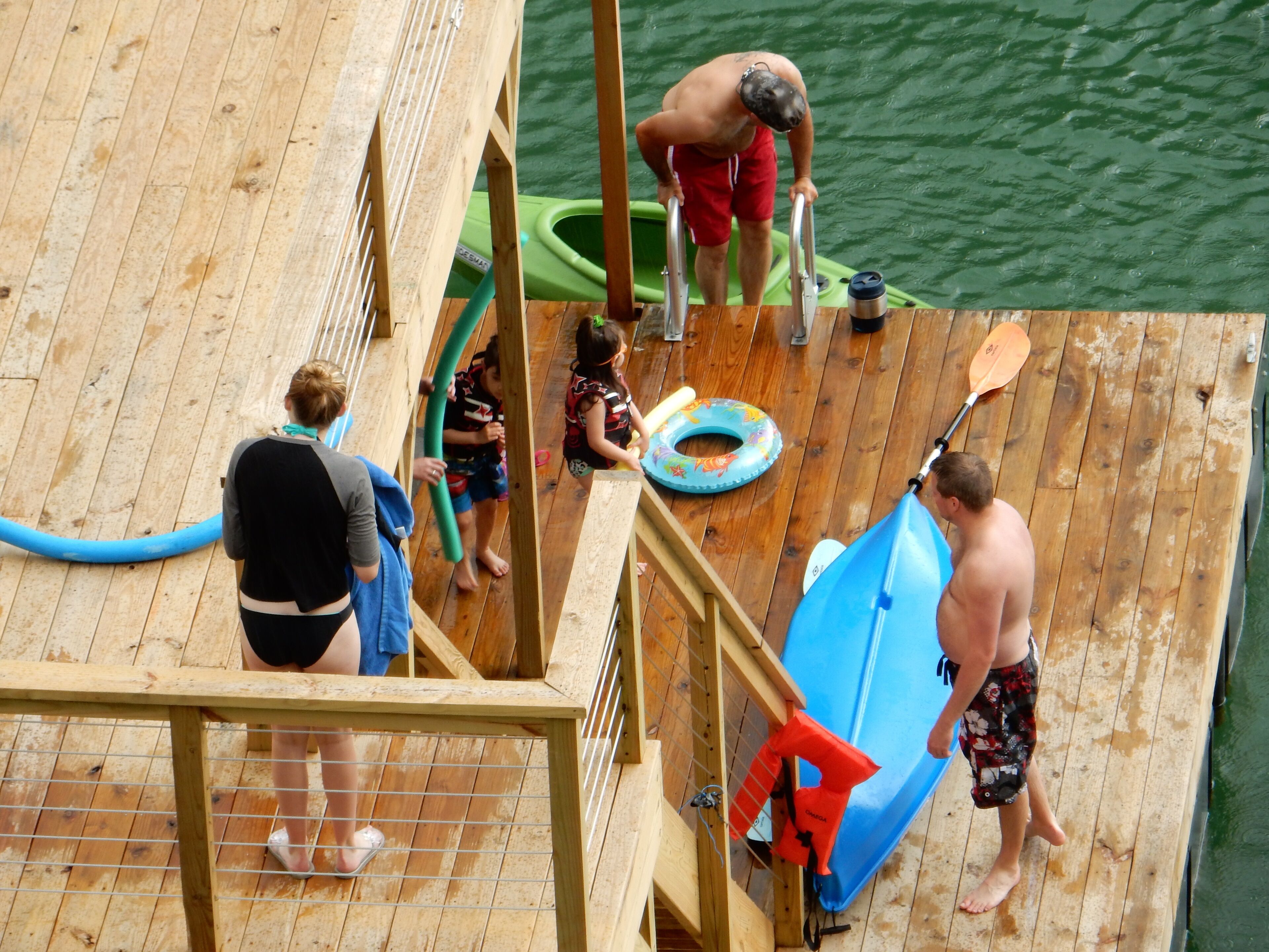 A wooden deck with a metal railing, a green kayak, a blue paddleboard, and a life preserver.