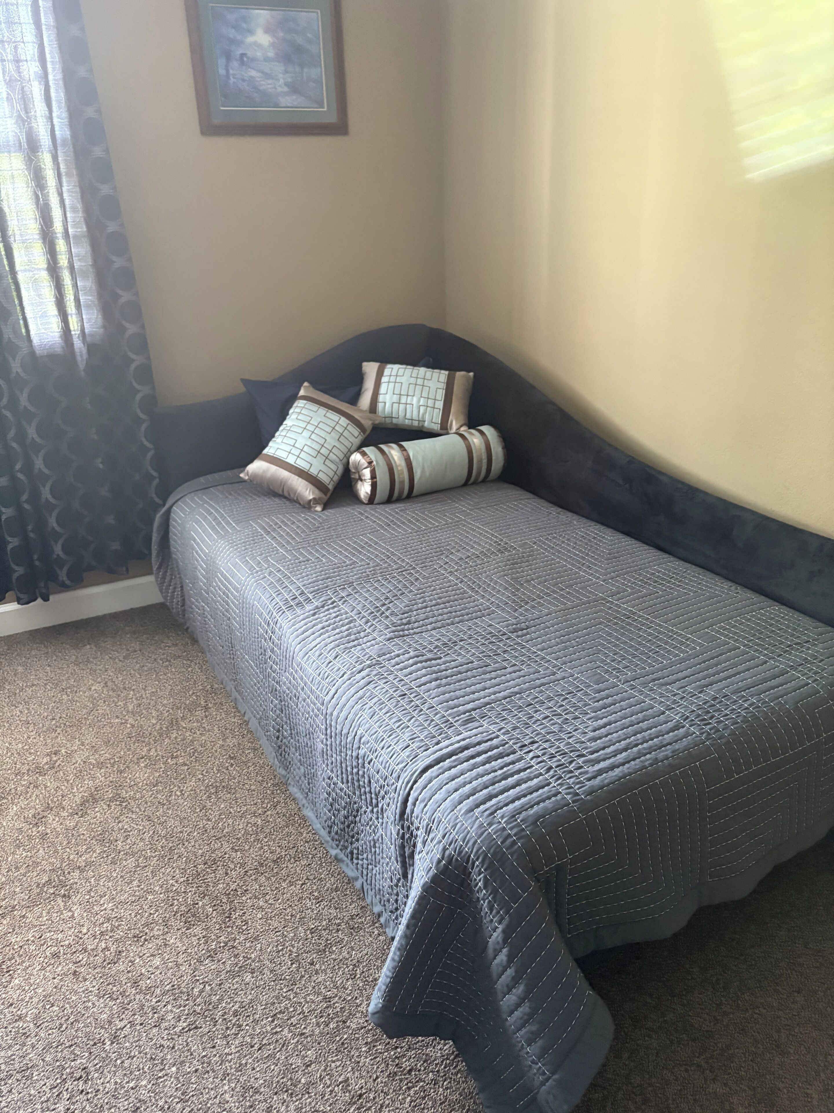 Ein Schlafzimmer mit einem Bett, einem dunklen Kopfteil, gemusterten Kissen, einem gerahmten Bild an der Wand und einem Vorhang mit Muster.