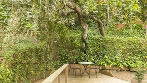 Une terrasse en bois avec une table et des chaises, entourée d’une végétation luxuriante et d’un arbre.