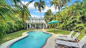 A swimming pool with a curved edge, surrounded by a brick deck. There are lounge chairs and a covered area with a green umbrella.