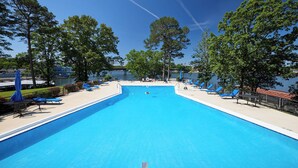 Ein rechteckiger Pool mit blauem Wasser, umgeben von Liegestühlen und Sonnenschirmen, mit Bäumen und Blick auf ein Gewässer im Hintergrund.