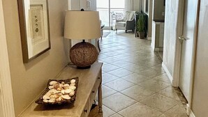 A hallway with a tiled floor, a console table with a lamp, a framed picture, and a basket. - Gulf front Beach Colony W w/seasonal beach chair/umbrella service March-October (Navarre Beach)